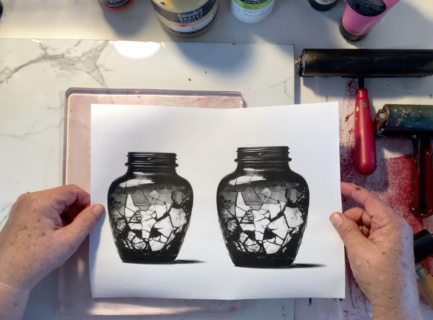 Two hands holding an image transfer print of two black glass jars with shattered glass design on a marble surface.
